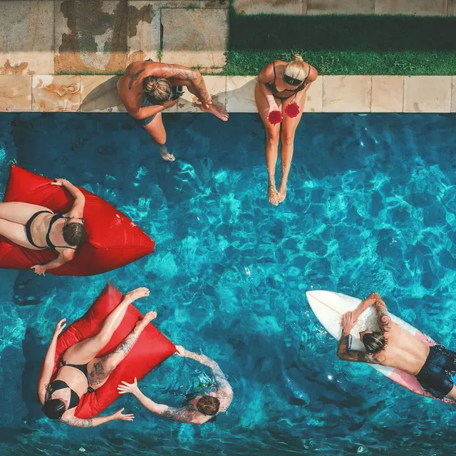 Aerial view of people enjoying a swimming pool. Some are on floaties, while others are sitting by the edge. Water is clear and inviting.