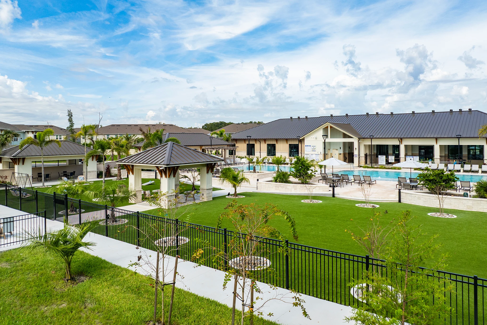 A spacious residential community featuring a swimming pool, lounging areas, palm trees, and buildings in the background under a blue sky with fluffy clouds.