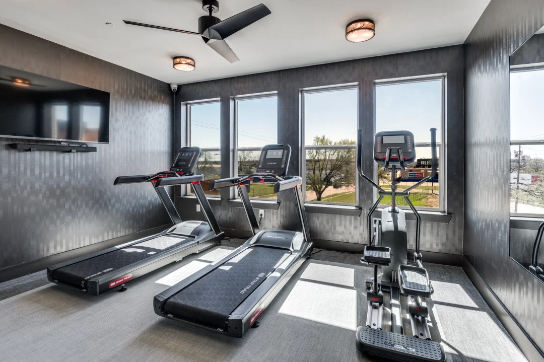 A modern fitness room with two treadmills and a cross trainer situated near large windows that provide natural light. The room has a contemporary design with dark walls, a ceiling fan, and a TV mounted on the wall.