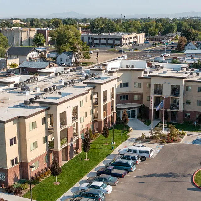 Aerial view of a modern residential complex with multiple buildings and parking areas, surrounded by greenery and city infrastructure.