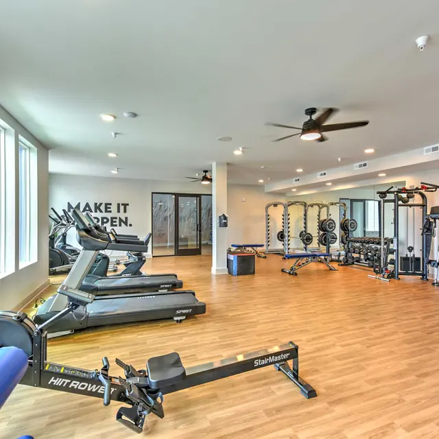 A spacious, modern gym with fitness equipment including treadmills, a rowing machine, and weightlifting stations. Natural light enters through large windows, and motivational text decorates the wall.