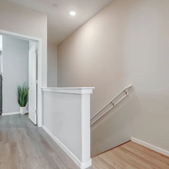A modern hallway featuring light walls, a wooden floor, and a staircase on the right, with a plant in the background.