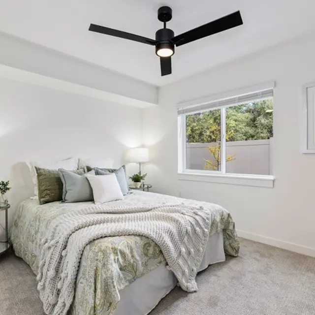 A well-decorated modern bedroom featuring a king-sized bed with a patterned comforter, two bedside lamps, a ceiling fan, and a window showing greenery outside.