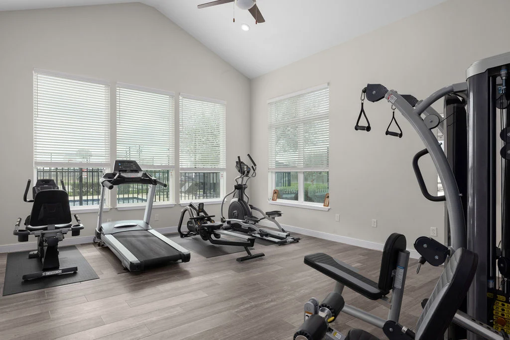 A spacious home gym featuring a treadmill, elliptical machine, and multi-station gym equipment. The room has large windows allowing natural light, and light-colored walls with wooden flooring.