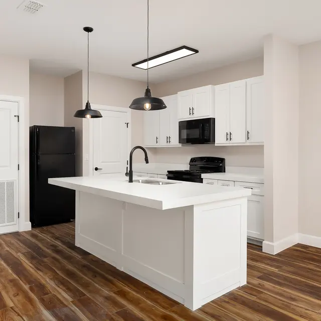 A spacious modern kitchen featuring a white island, dark pendant lights, and wooden flooring. Appliances include a black refrigerator and a microwave surrounded by white cabinetry.