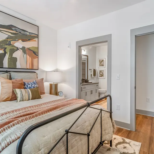 A cozy bedroom featuring a bed with a patterned blanket, decorative pillows, and a large painting above it. There's a nightstand with a lamp and a glimpse of an attached bathroom area in the background.