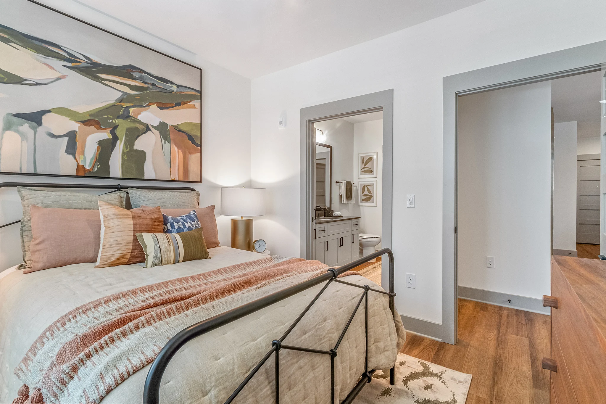 Contemporary Cozy Bedroom A cozy bedroom featuring a bed with a patterned blanket, decorative pillows, and a large painting above it. There's a nightstand with a lamp and a glimpse of an attached bathroom area in the background.