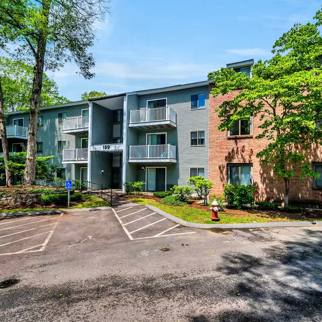 A view of a multi-story apartment building surrounded by trees and green landscaping. The building features balconies and a mix of brick and grey exterior. There are designated parking spaces in front.