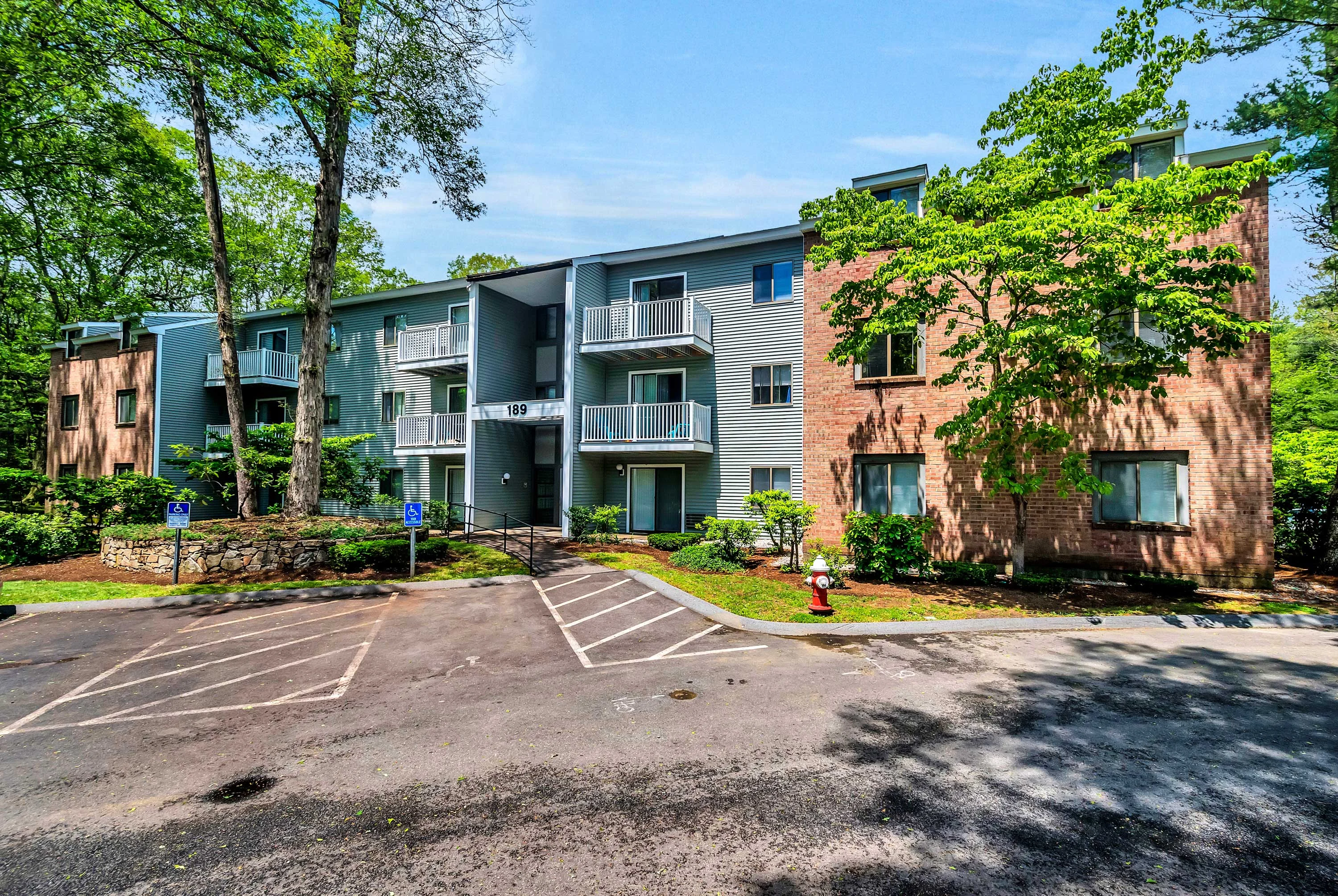 Apartment Complex Exterior View A view of a multi-story apartment building surrounded by trees and green landscaping. The building features balconies and a mix of brick and grey exterior. There are designated parking spaces in front.