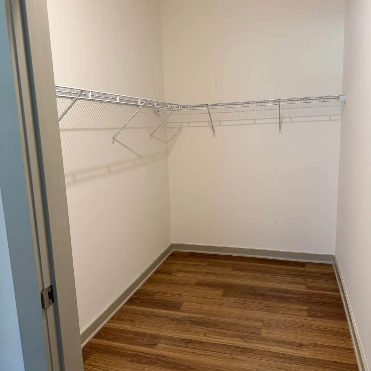 An empty closet with white walls and a wooden floor, featuring a wire shelving unit along one wall.
