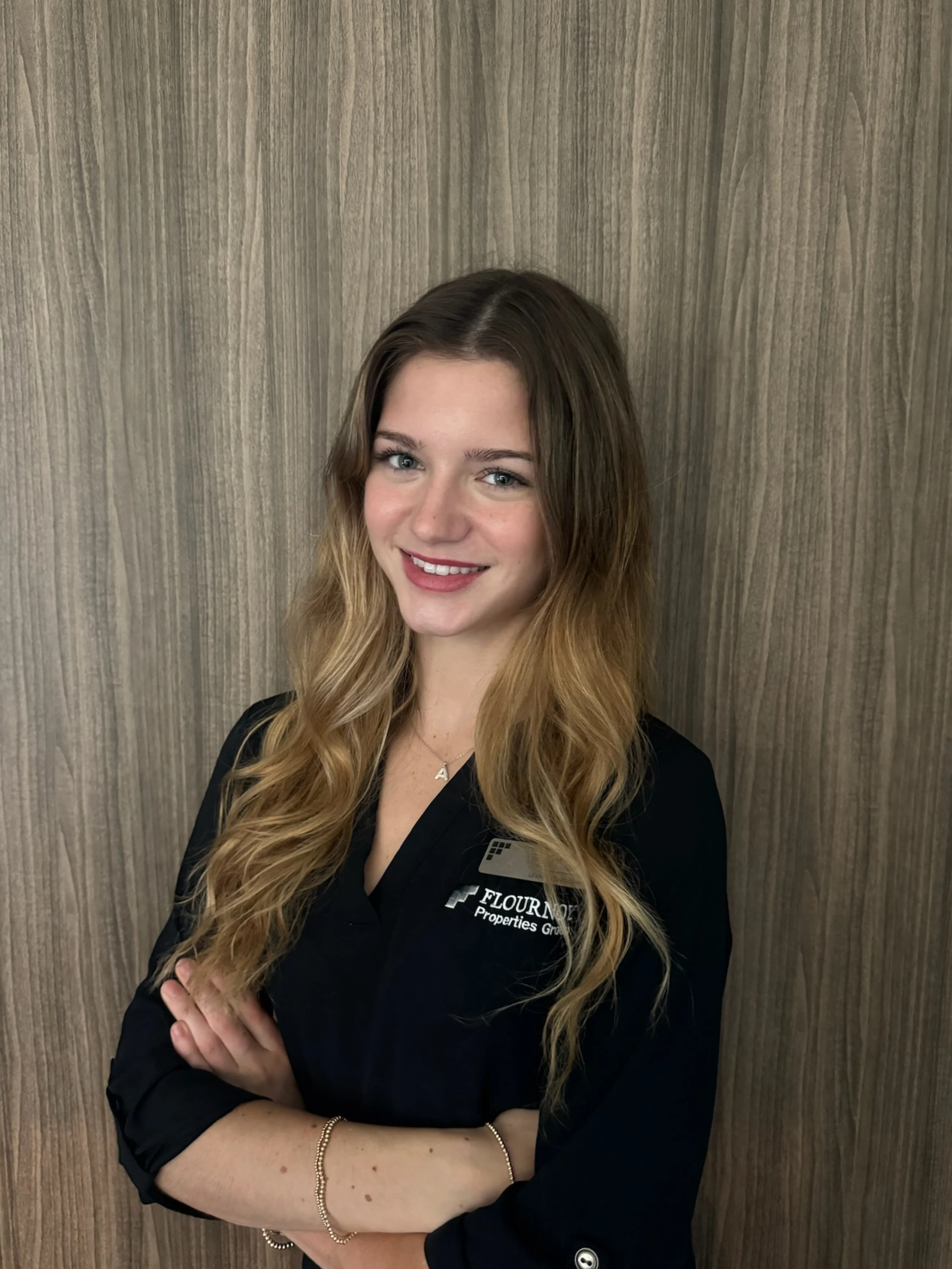 Professional Portrait of a Woman A young woman with long, wavy dark blonde hair wearing a black professional shirt, smiling and posing with her arms crossed against a wooden textured background.