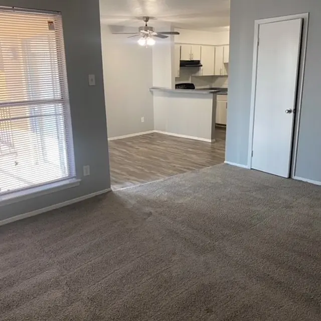 A bright living room with carpet flooring and a view of a kitchen in the background. The room features a ceiling fan and a window with blinds through which natural light enters.