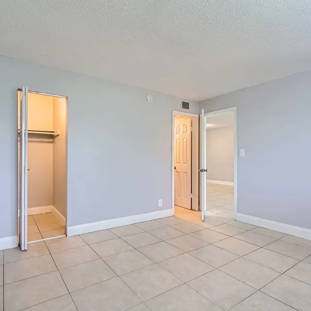 An empty room featuring light gray walls, a tiled floor, and a closet on the left with an open door. The right side shows a door leading to another room.