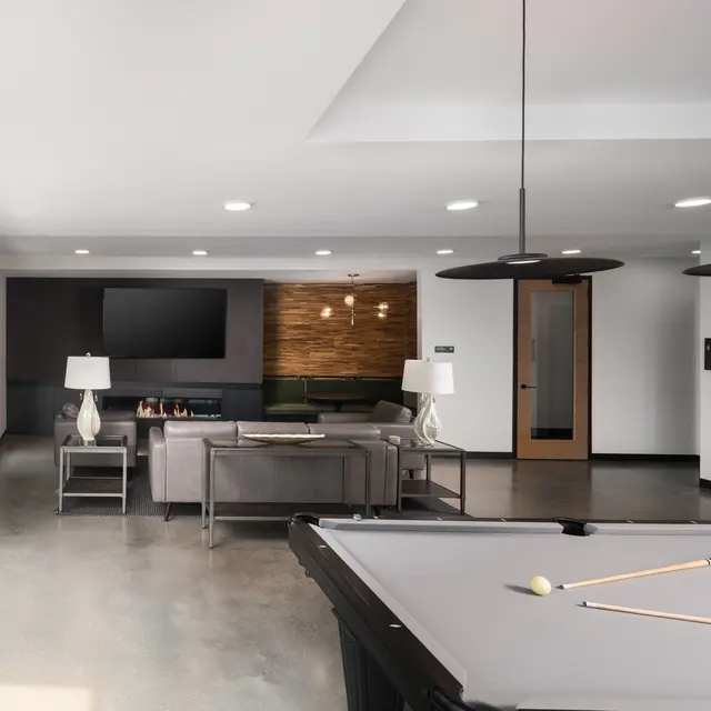 A spacious modern living area featuring a billiard table, minimalist furniture, and large windows allowing natural light. The room has a contemporary design with a TV mounted on the wall and stylish lamps on side tables.