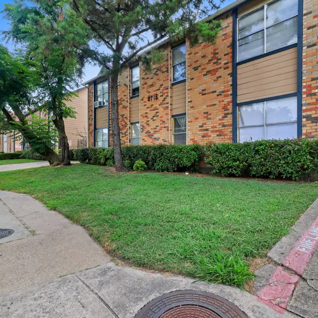 Exterior view of a two-story apartment building with brick and wood paneling, grassy area in front, and trees nearby.