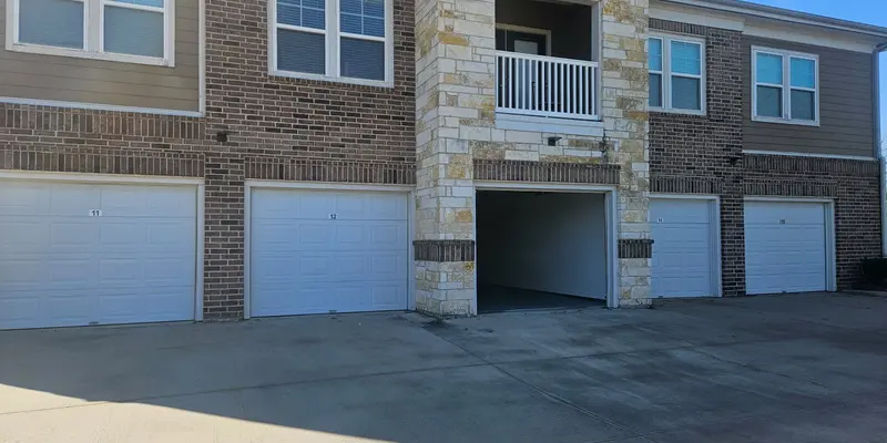 A two-story apartment complex featuring a stone and brick facade, with three garage doors on the ground level and a balcony above the entrance.