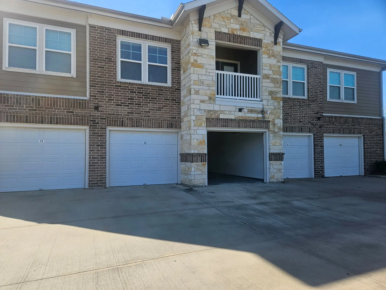 Apartment Complex A two-story apartment complex featuring a stone and brick facade, with three garage doors on the ground level and a balcony above the entrance.