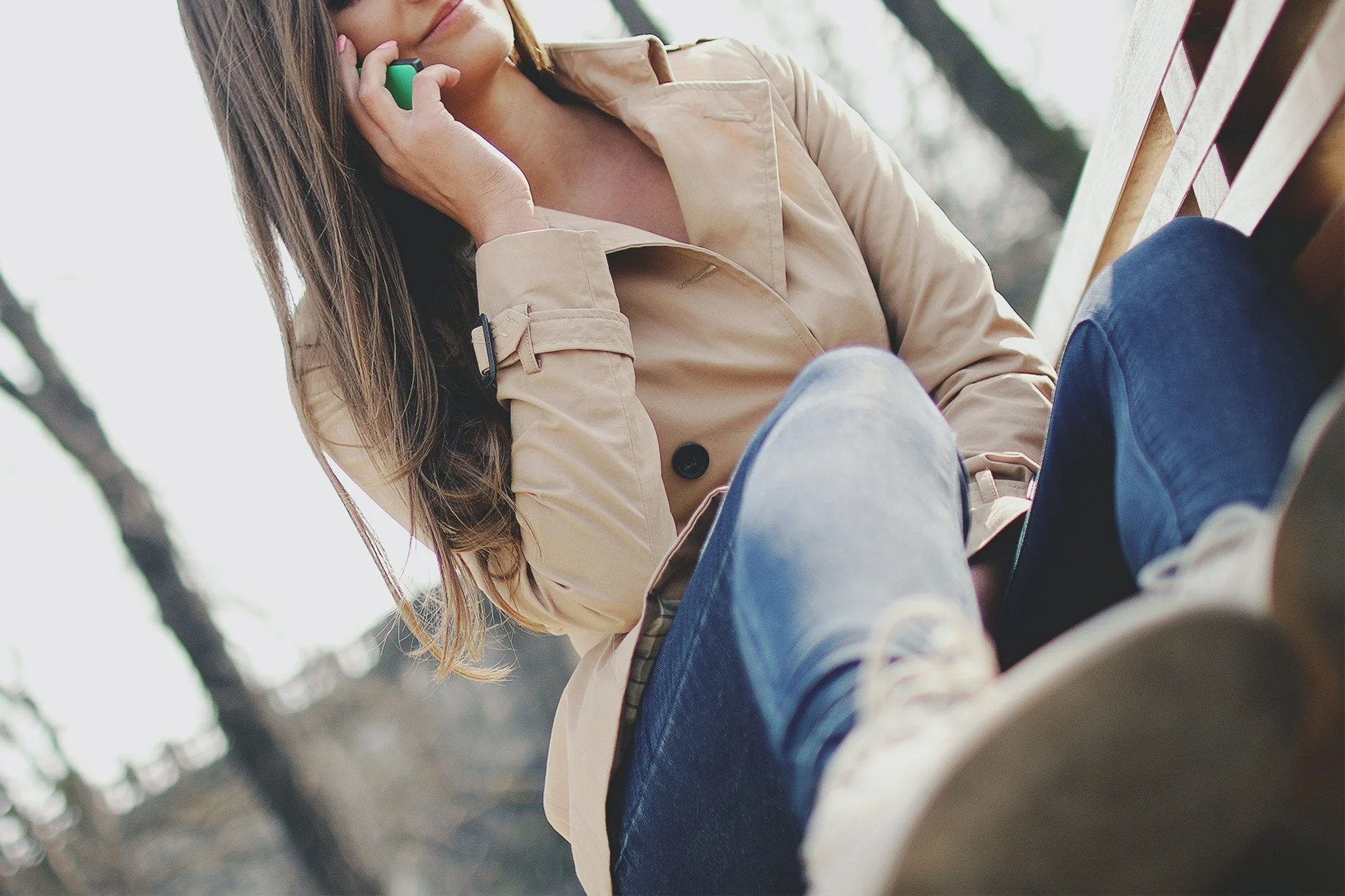 A woman sitting on a bench outdoors, dressed in a beige trench coat and blue jeans. She has long, wavy hair and is holding a smartphone to her ear, while looking off to the side thoughtfully.