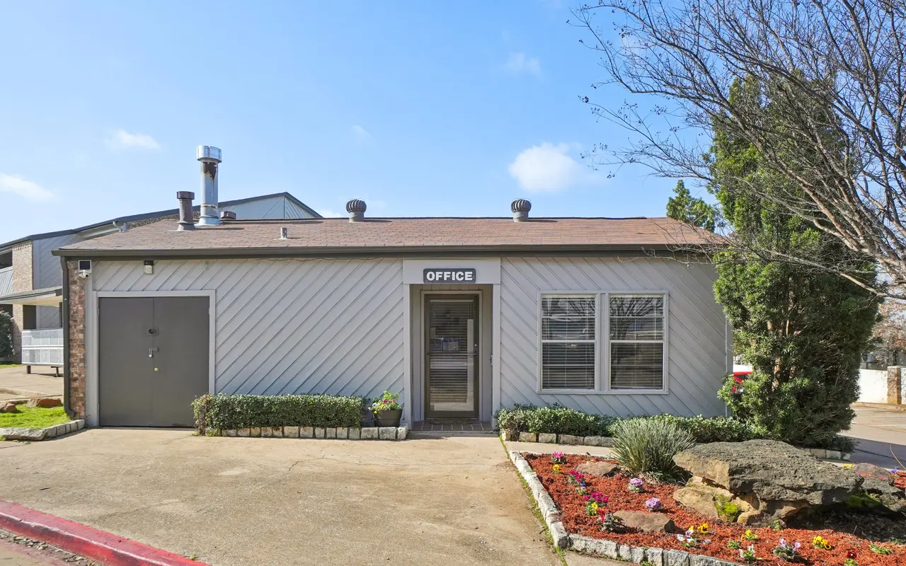 Exterior view of a one-story office building with a sign that reads 'OFFICE'. The building features beige siding, multiple windows, and a small landscaped area in front. A parking area is visible to the left.