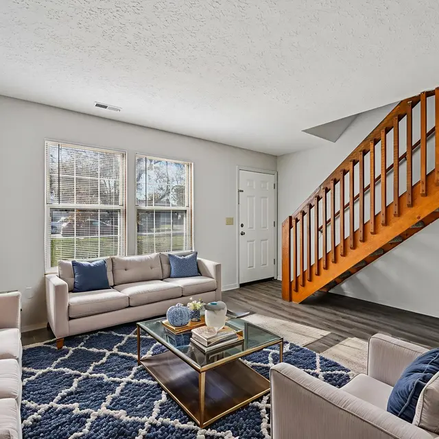 A modern living room with two beige couches and a blue rug. There is a wooden staircase in the background and large windows providing natural light.