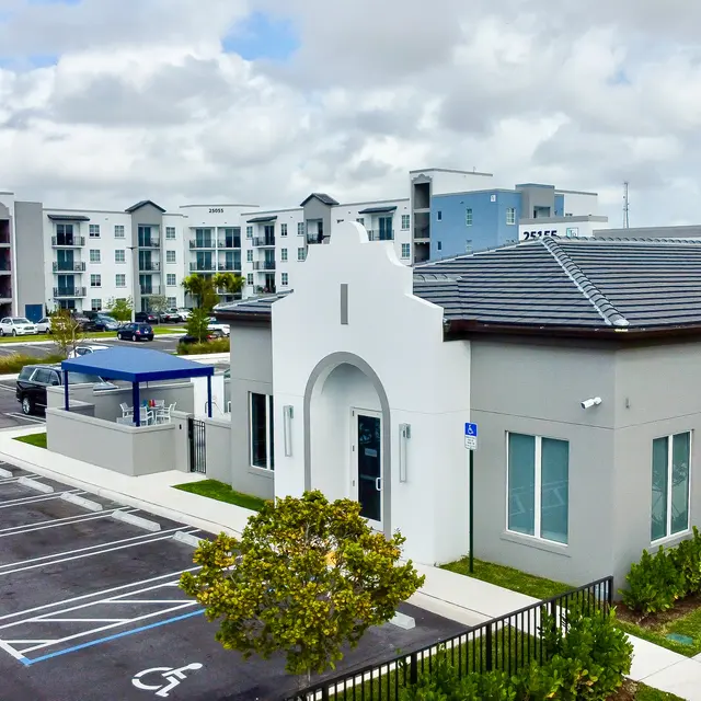 Modern Building with Parking Lot A modern building with a unique architectural design, surrounded by a parking lot and greenery. In the background, there are multiple apartment buildings.
