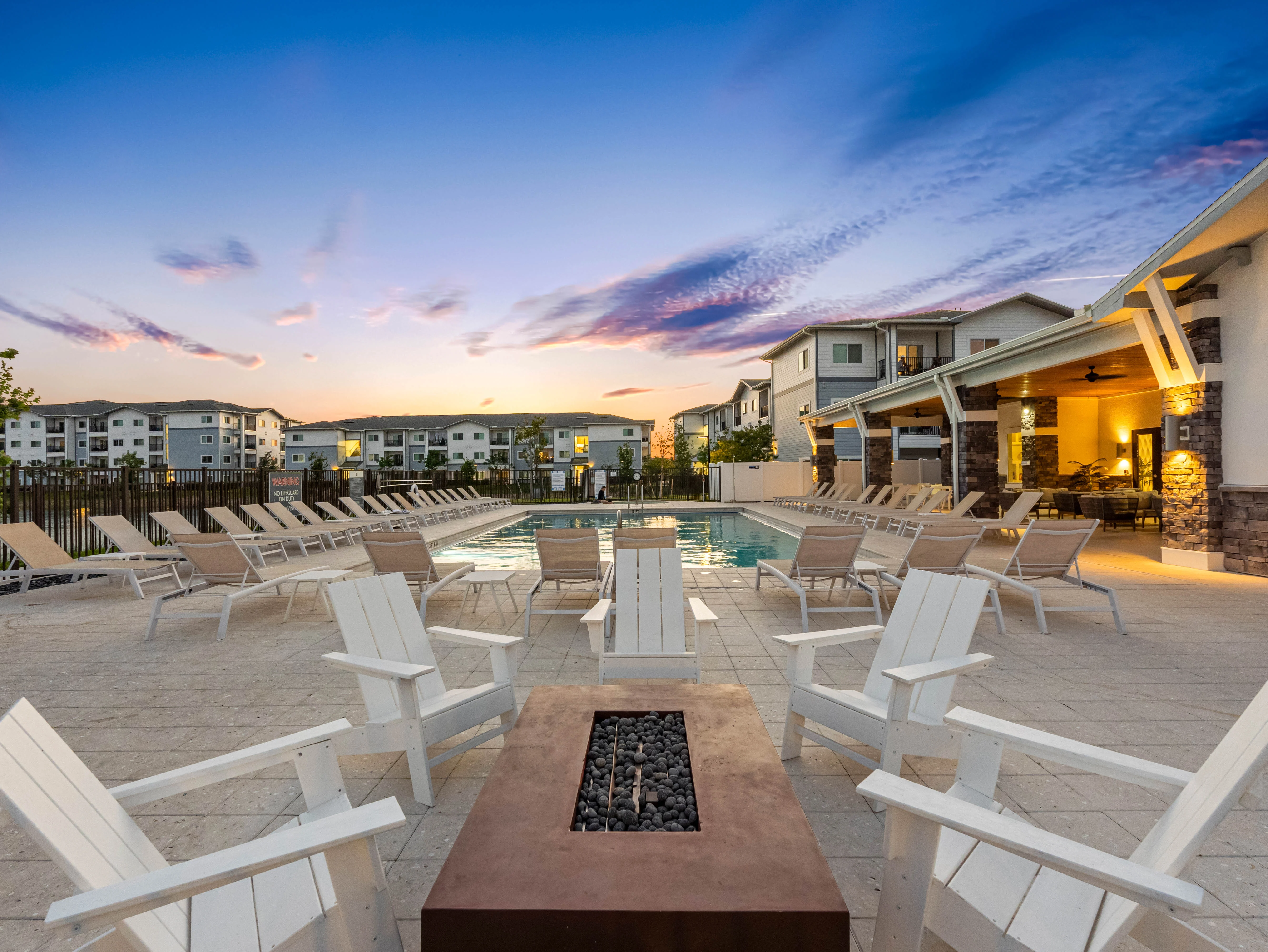 A serene poolside area during twilight with lounge chairs arranged around a fire pit. The sky features a gradient of blue and pink hues, and residential buildings are visible in the background.