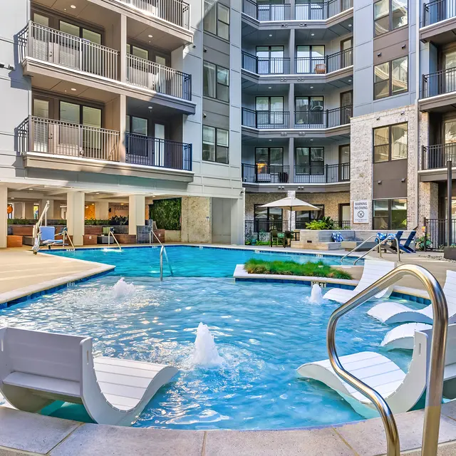 An outdoor pool area in a modern apartment complex featuring lounge chairs, umbrellas, and water fountains.