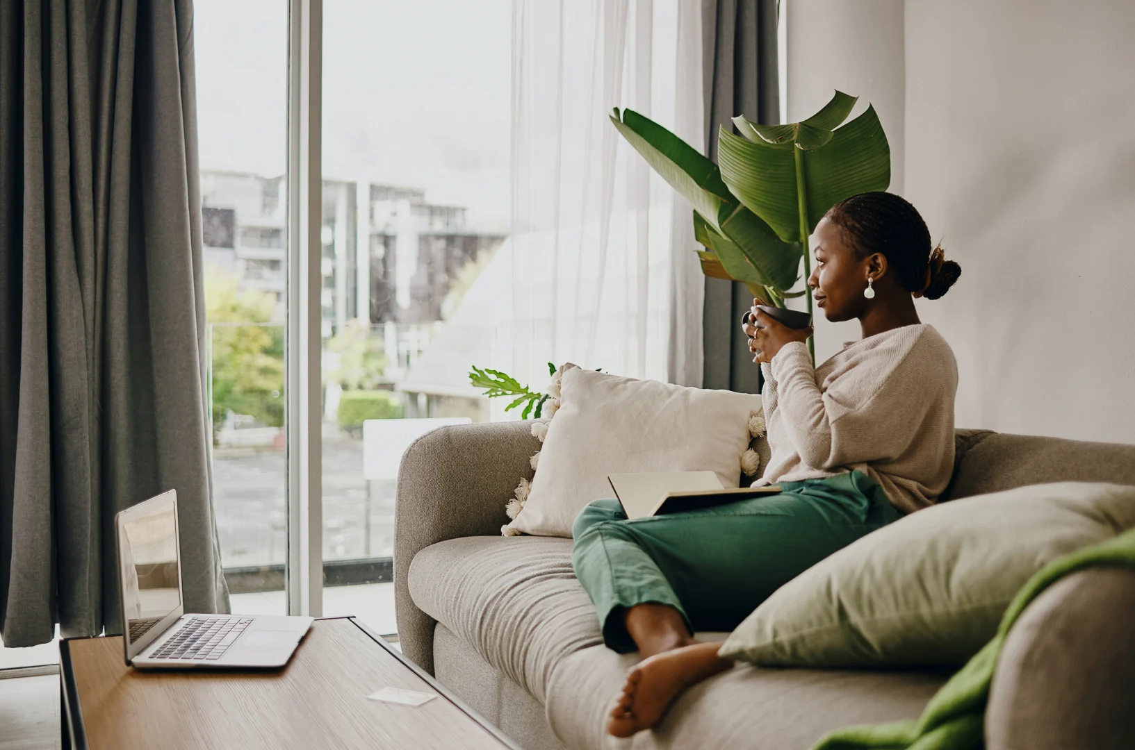 Relaxing in a Cozy Living Room A woman sitting on a sofa with a plant in her hands, surrounded by a cozy living room atmosphere. She is looking out the window, with a laptop on a table nearby.