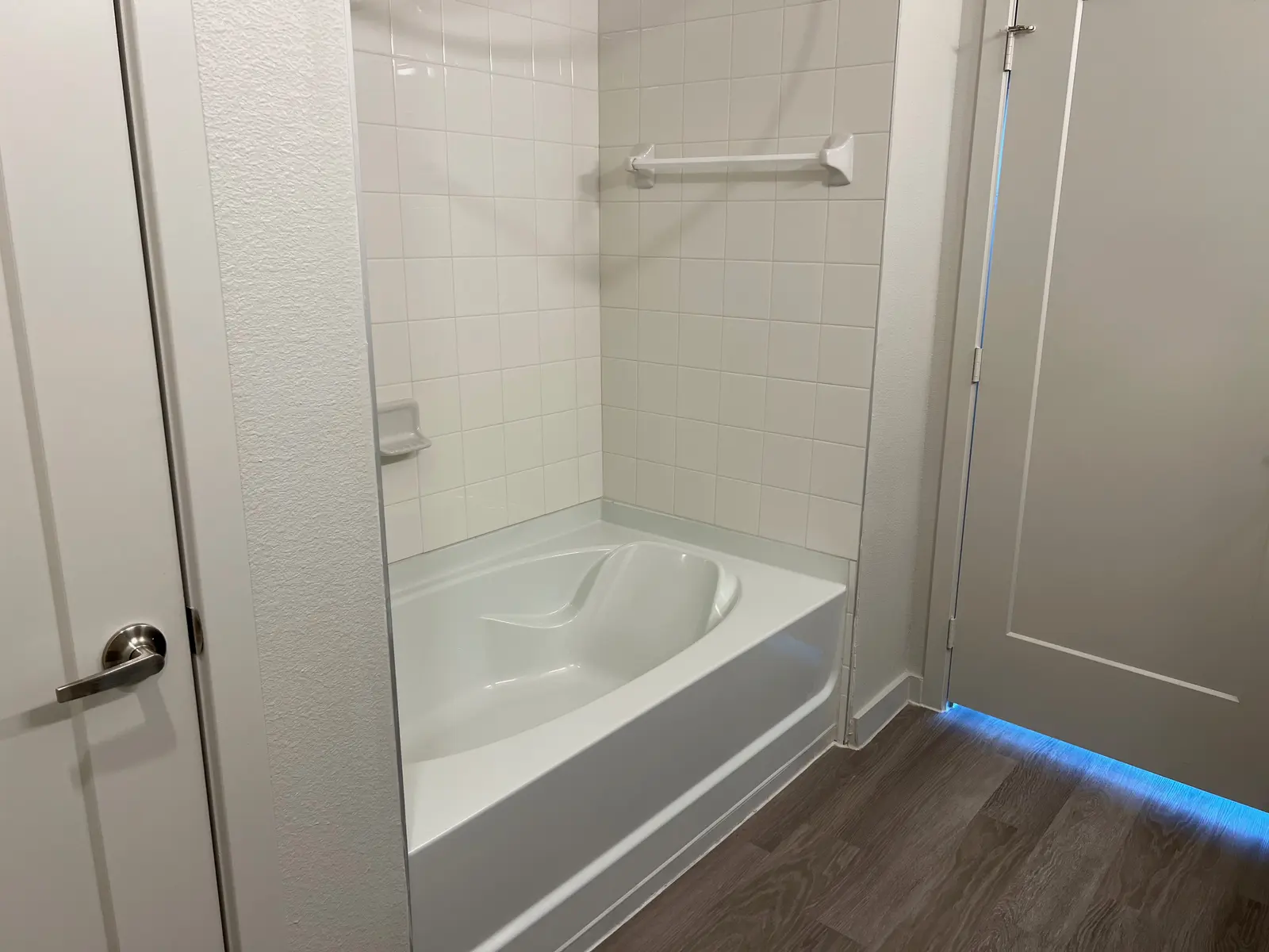 A modern bathroom featuring a white bathtub next to a wall with white tiles and a ceiling-mounted grab bar. The floor is wooden, with a door visible in the background.