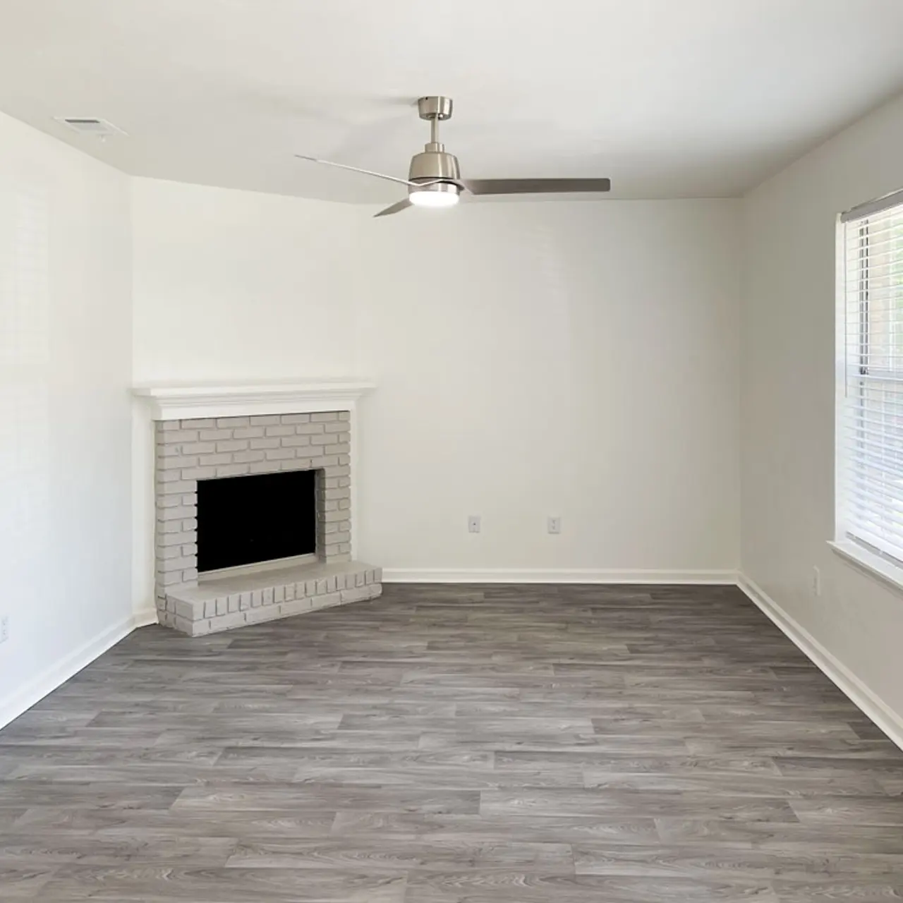 Spacious living room with a white brick fireplace, light gray wooden flooring, and large windows with white blinds.