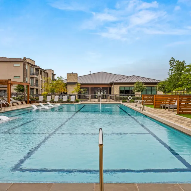 A large swimming pool with lounge chairs on the side, surrounded by modern apartment buildings and greenery, under a clear blue sky.