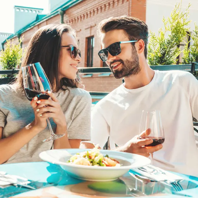 A couple sitting at a table on a terrace, enjoying drinks and a meal. The woman holds a glass of red wine and smiles at the man, who wears sunglasses and has a glass of wine in front of him. There is a bowl of salad on the table and greenery in the background.