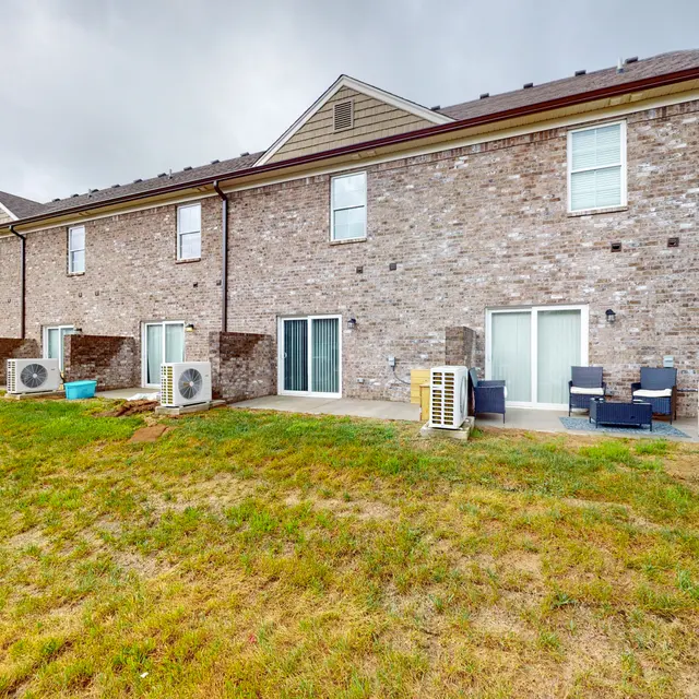 Back view of brick apartment units with outdoor seating and air conditioning units, featuring a grassy area.