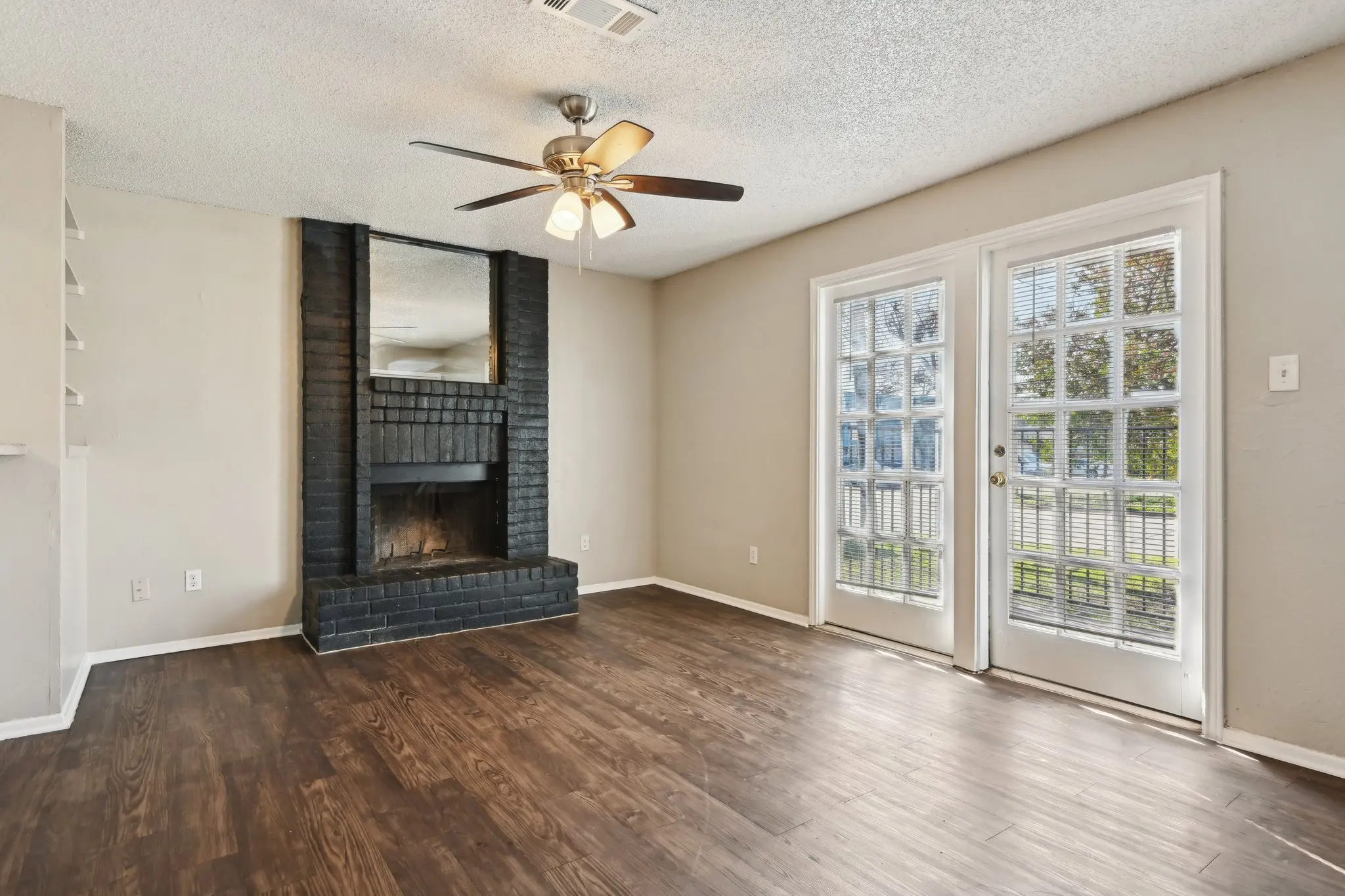 A spacious living room featuring hardwood flooring, a ceiling fan, a black brick fireplace, and double doors leading outside.