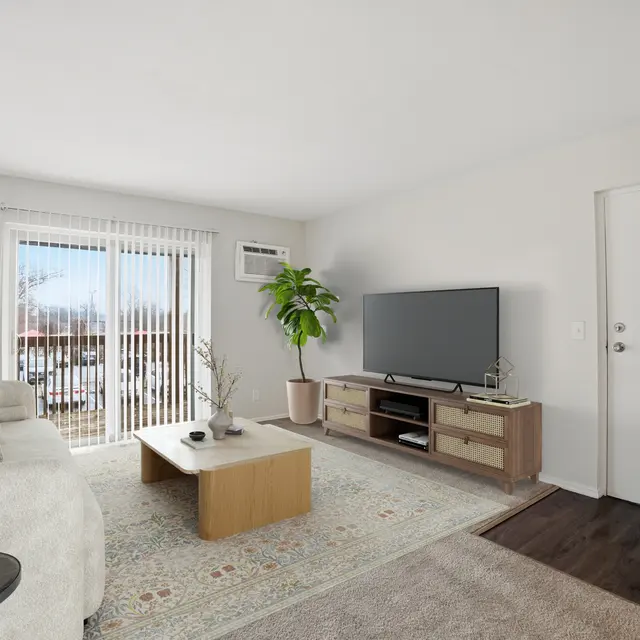 A modern living room featuring a light-colored sofa, a wooden coffee table, and a television on a cabinet. Large windows provide natural light.