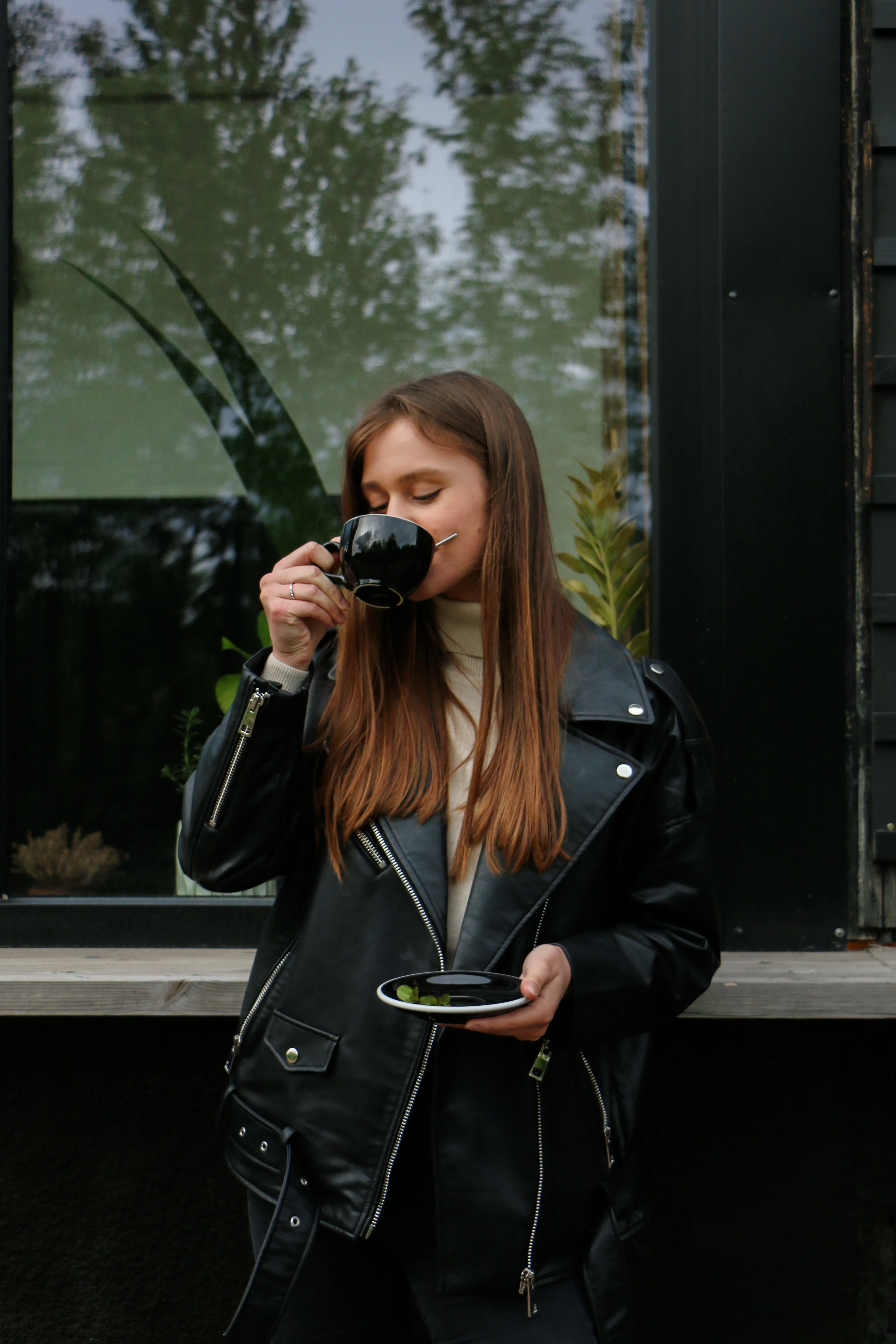 Coffee Break A woman in a black leather jacket drinks from a black cup while holding a plate with a small item, standing in front of a window adorned with greenery.