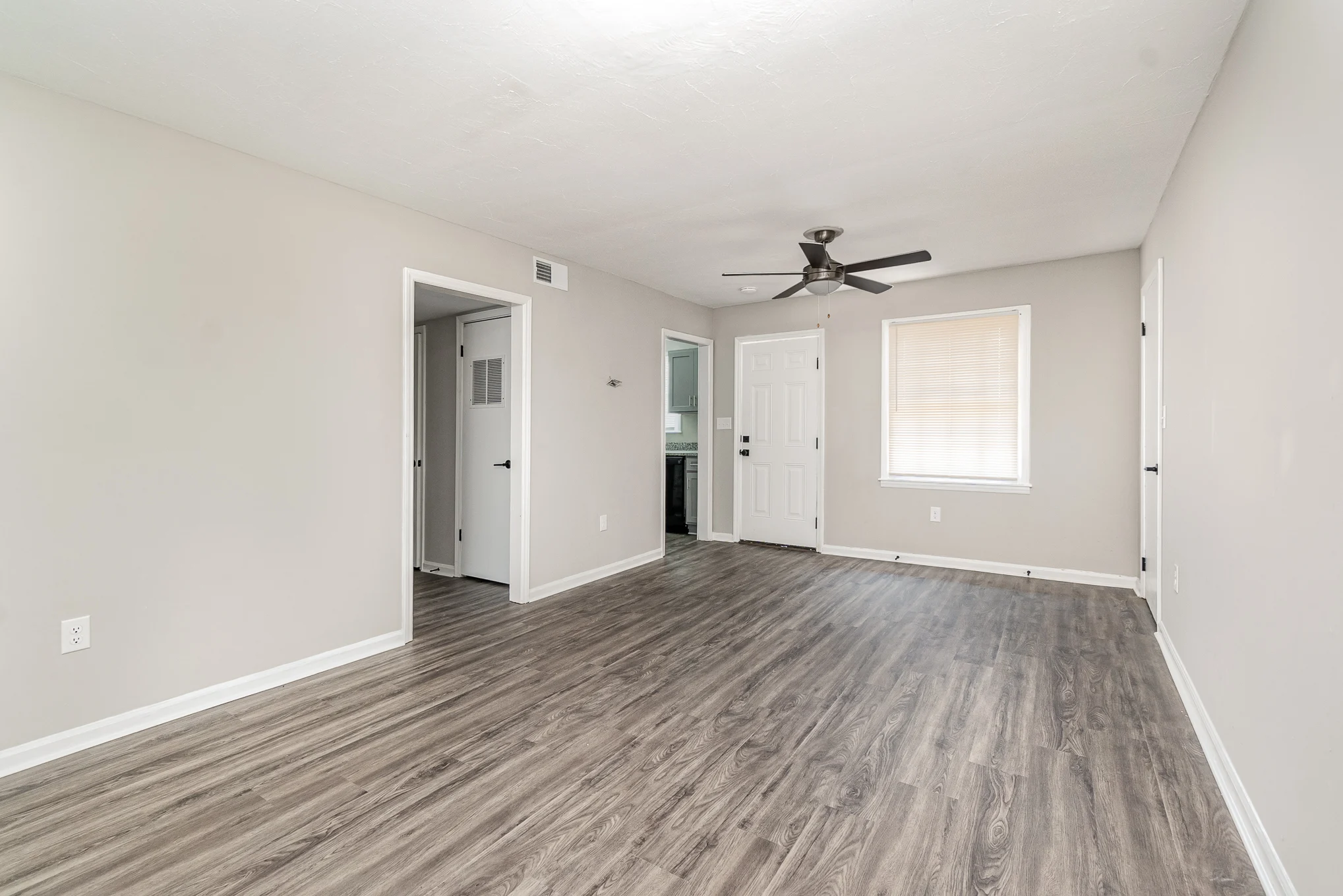 A spacious, neutral-toned living room with light wood flooring, a ceiling fan, and two doors leading to other rooms.