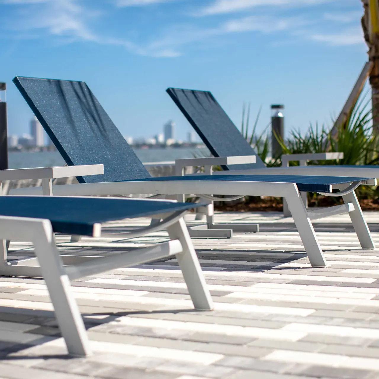 Three modern lounge chairs positioned along a waterfront patio with a view of the city skyline in the background.