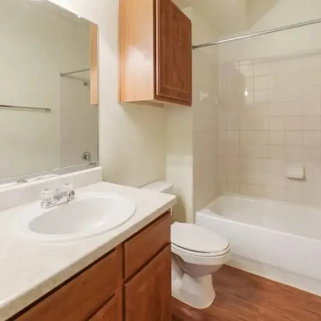 A clean and simple bathroom featuring a bathtub, toilet, and a sink with a mirror above it. The cabinetry is wooden, and the floor is wooden laminate.