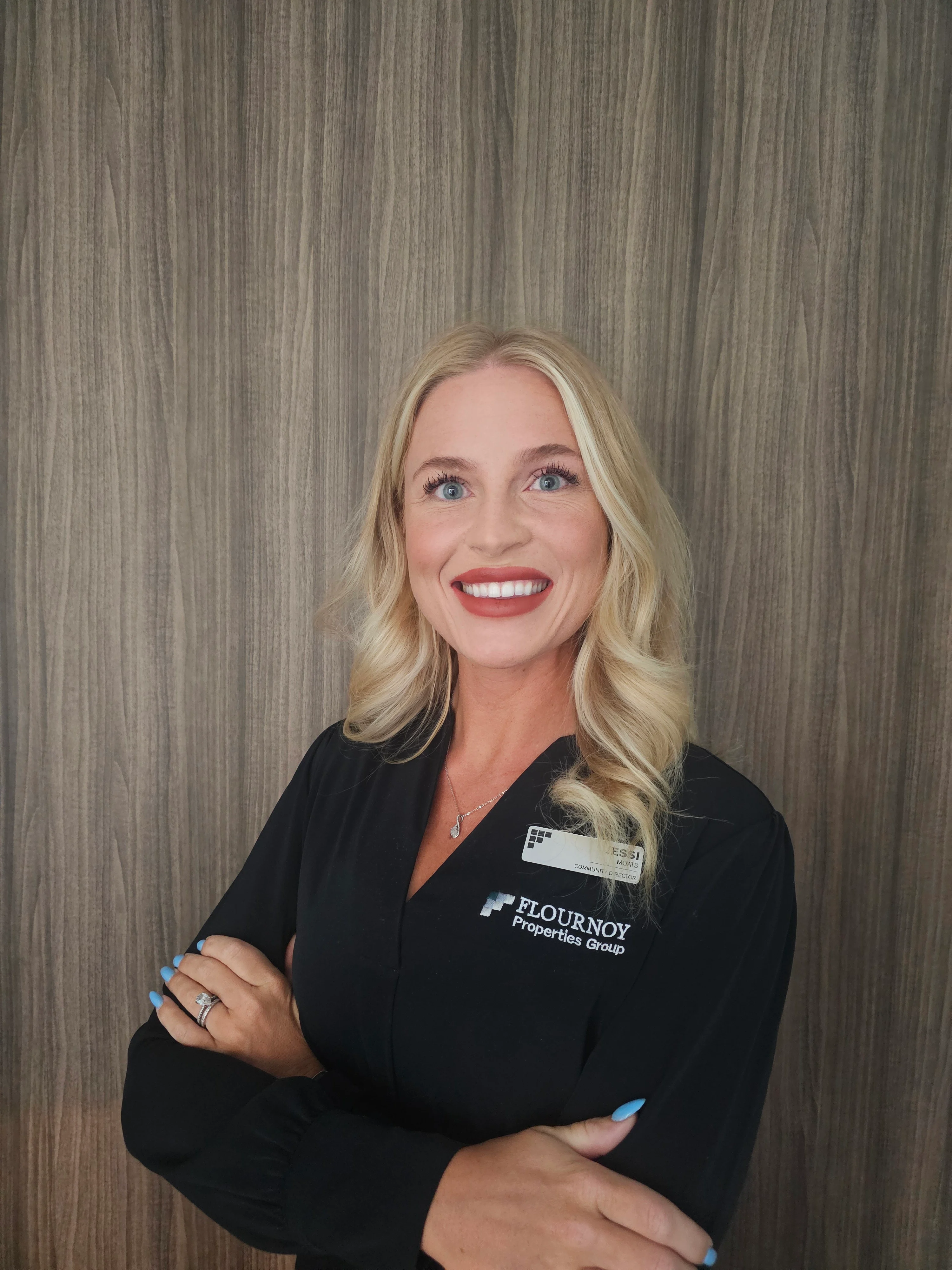 Professional Portrait of Flournoy Property Group Representative A smiling woman with blonde hair standing with her arms crossed in front of a wooden wall. She is wearing a black shirt with a name tag from Flournoy Property Group.