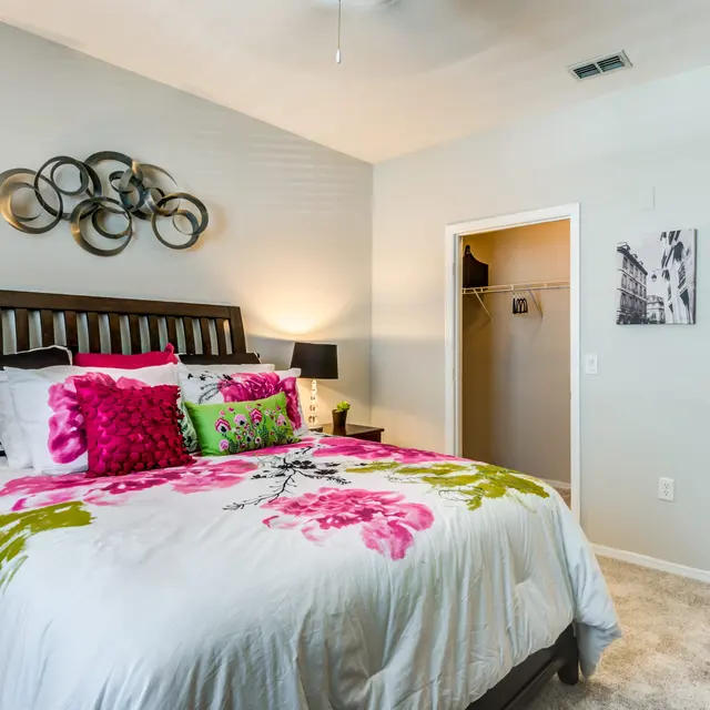 A cozy bedroom with a large bed adorned with colorful pillows and a floral bedding set. There are bedside lamps on either side and a wall decoration above the headboard. An open closet is visible to the left, and a door leading to another room is on the right.