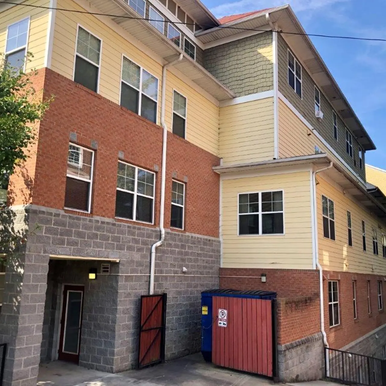 A multi-story student housing building with a combination of brick and siding exteriors, surrounded by greenery and a blue dumpster in the foreground.