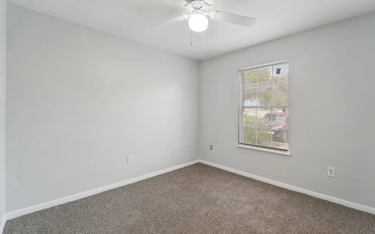 An empty bedroom with light gray walls and carpeted floor. A ceiling fan is visible, and there is a window with a slight view of the outdoors.