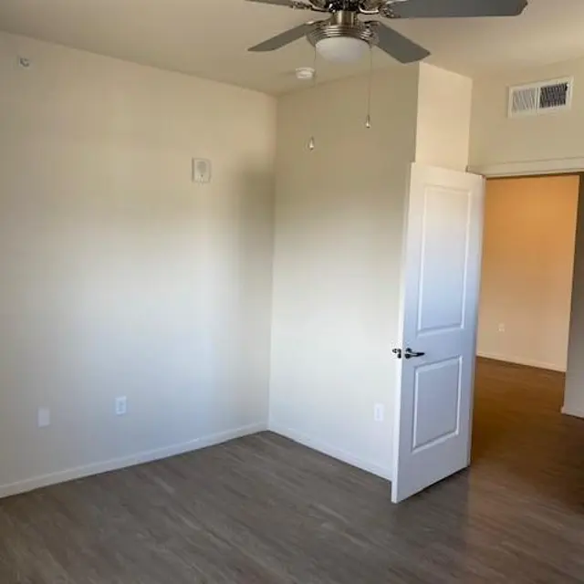 An empty room with light-colored walls, a ceiling fan, and laminate flooring. A closed door leads to another room in the background.
