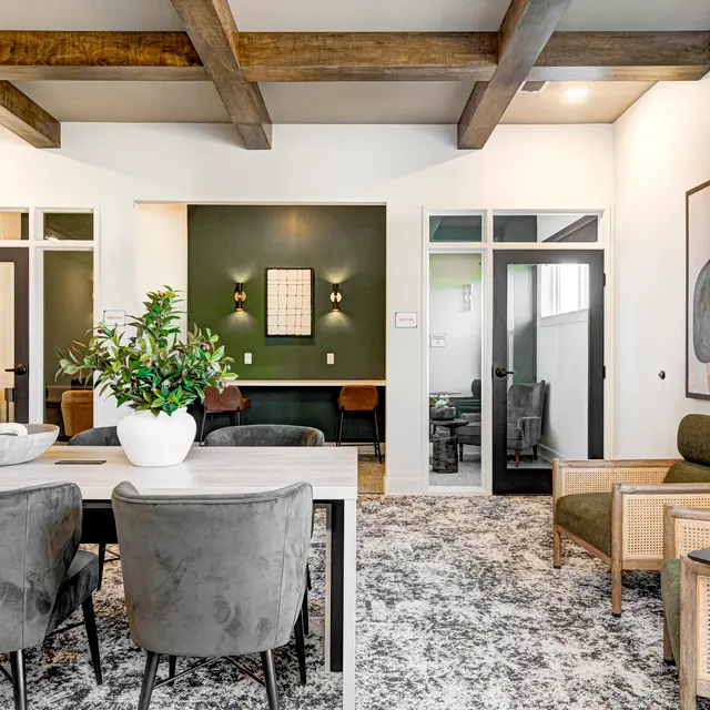 A modern living space featuring a dining table with chairs, a green accent wall, and stylish furniture including a lounge chair. The area is well-lit with wooden beams on the ceiling.