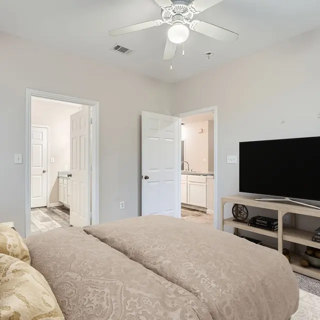 A cozy bedroom featuring a large bed covered with a patterned duvet, a nightstand with a lamp, and a modern television on a wooden stand. Two doors lead to other rooms, providing a glimpse of a bathroom and possibly another room.