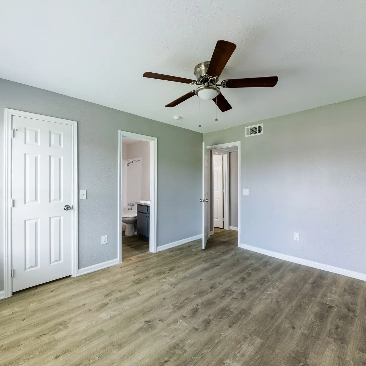 A spacious modern bedroom featuring a ceiling fan, light gray walls, and light wood flooring. There are two white doors leading to other rooms, and natural light coming in through a window.