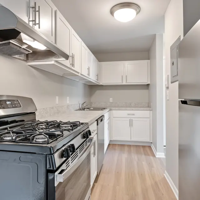 A modern kitchen featuring white cabinetry, a stainless steel gas stove, and a silver refrigerator. The countertop is light-colored with a simple design, and the floor has a wooden appearance.