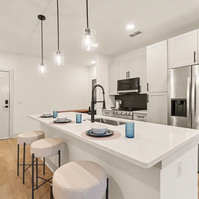 A modern kitchen featuring a white island with beige stools, three hanging light fixtures, and stainless steel appliances.