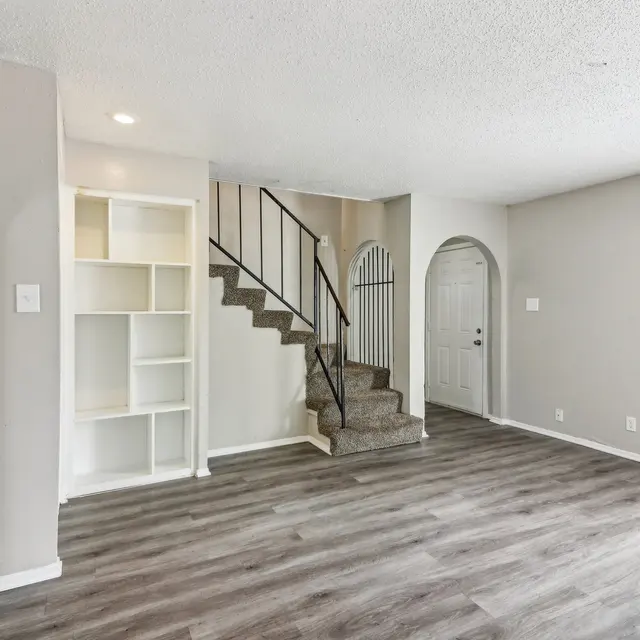 A spacious living room featuring light wooden flooring, a staircase leading to an upper level, a built-in bookshelf on the left, and an archway leading to another room. The walls are painted in a neutral tone, and there is an entrance door visible in the background.